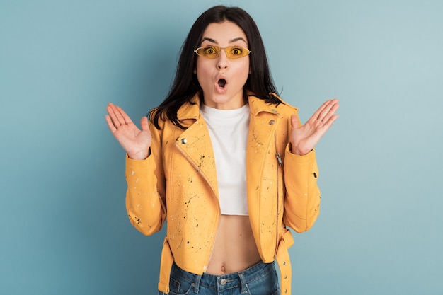 Surprised, charming woman in sunglasses and orange jacket on a blue wall