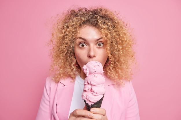 Surprised curly haired young woman licks ice cream eats junk food eats appetizing summer dessert dressed in stylish outfit poses against pink wall. Lovely European female bites sweet snack