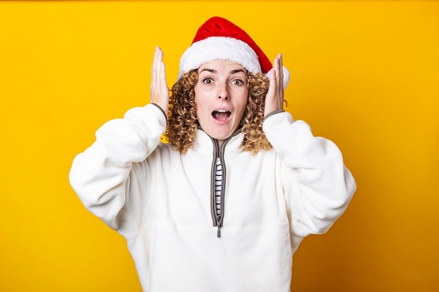 Surprised curly young woman in santa hat on a yellow background.