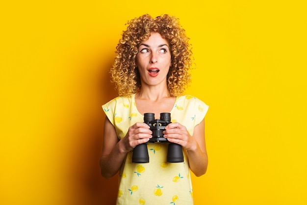 Surprised curly young woman with binoculars on yellow surface