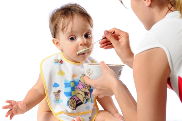 Surprised cute little baby girl eating food from spoon Mother holding plate and feeding child Isolated on white background