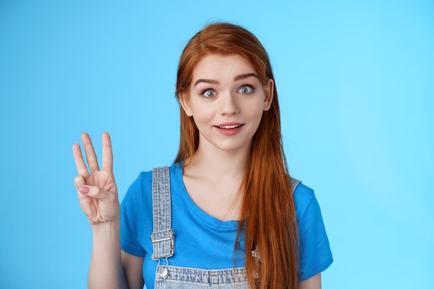 Surprised cute redhead caucasian girl show number three, third, counting fingers, raise eyebrows ambushed, smiling curiously, stand blue background playful tender expression