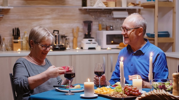 Surprised elderly woman with charming smile sitting at the table in kitchen opening small gift box present. Cheerful couple dining together at home, enjoying the meal, celebrating their anniversary