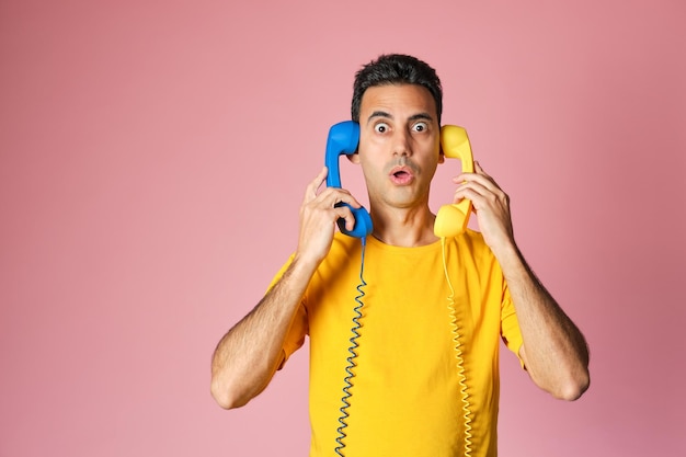 Surprised face brunette boy in yellow tshirt holding two vintage blue and yellow phones in ears on pink background