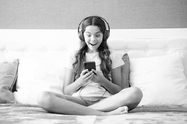 Surprised face surprise emotions of teenager girl Teenager child girl wearing headphones watching videos listening music on smart phone sitting on bed in her room