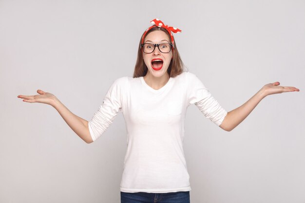 Surprised face with raised arms, portrait of beautiful emotional young woman in white t-shirt with freckles, black glasses, red lips and head band. indoor shot, isolated on light gray background.