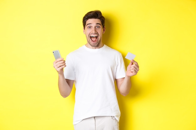 Surprised guy holding smartphone and credit card, online shopping on black friday, standing over yellow background