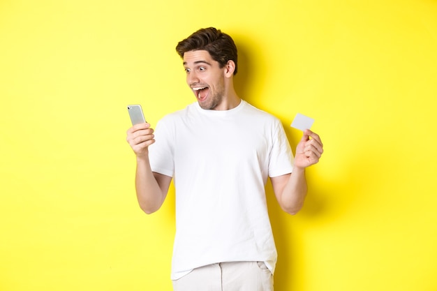 Surprised guy holding smartphone and credit card, online shopping on black friday, standing over yellow wall