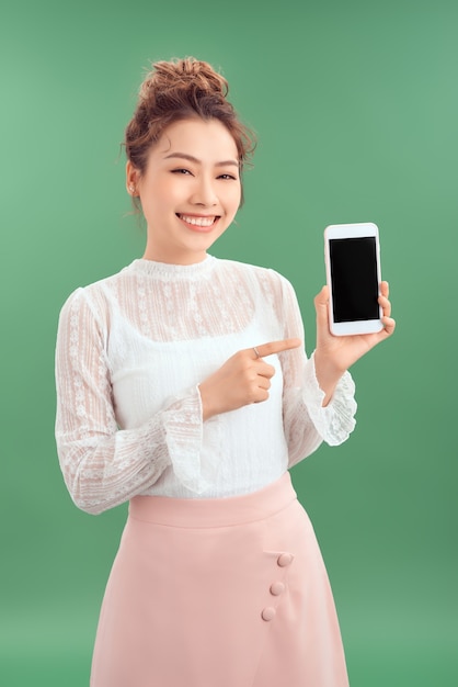 Surprised happy Asian woman showing blank smartphone screen and pointing on it. Isolated green background.