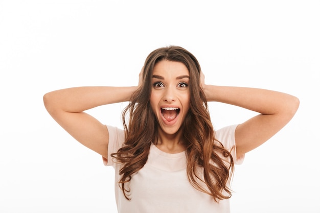 Surprised happy brunette woman in t-shirt screaming and covering her ears while over white wall