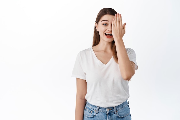 Surprised and happy girl covers half of face smiling and gasping amazed checking out something awesome standing in casual clothes against white background