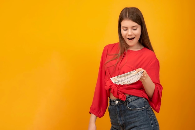 Surprised and happy teenage girl keeps wad of money Earnings for young people Yellow background Copy space