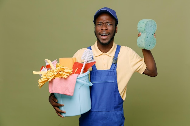 surprised holding bucket of cleaning tools with sponge young africanamerican cleaner male in uniform with gloves isolated on green background