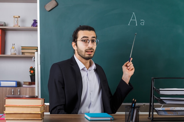 Surprised male teacher wearing glasses points at blackboard with pointer stick sitting at table with school tools in classroom