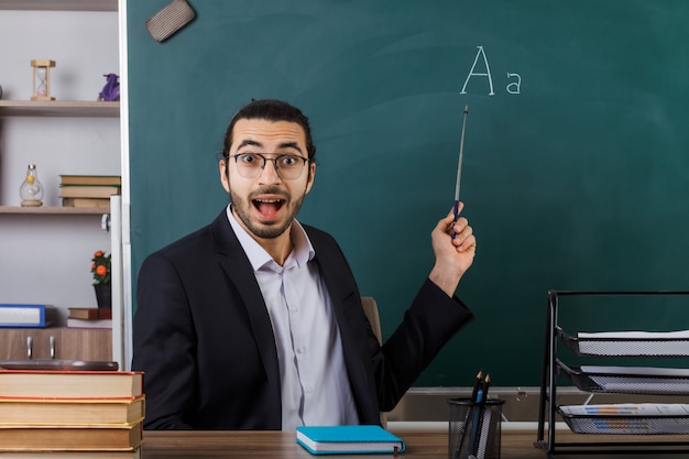 Surprised male teacher wearing glasses points at blackboard with pointer stick sitting at table with school tools in classroom