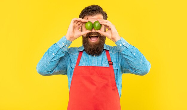 Surprised man in apron holding fresh limes on eyes yellow background, fruit seller.