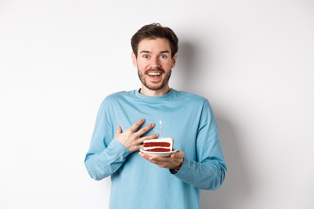 Surprised man receiving birthday cake with lit candle, being congratulated with b-day, gasping amazed, standing over white background.