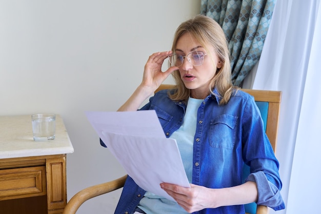 Surprised middle aged woman with glasses reading papers sitting at home in chair