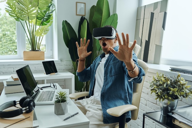 Surprised mixed race man wearing virtual reality glasses and gesturing while sitting at the desk