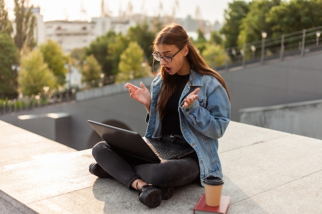 Surprised modern student woman in a denim jacket sitting on stairs look at screen laptop. Distance learning. Modern youth concept.