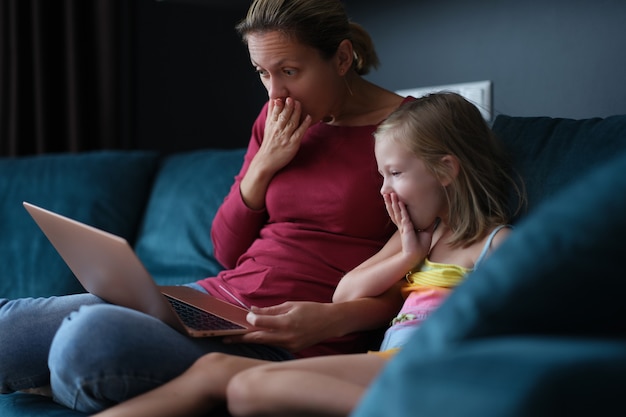 Surprised mom and daughter are sitting on couch and looking at laptop screen