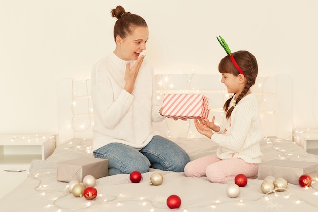 Surprised mother and daughter wearing casual style white sweaters sitting on bed, child giving present with love, astonished male take box and covering mouth with palm.