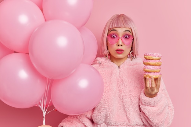 Surprised pink haired young Asian woman looks surprisingly wears fur coat holds pile of delicious doughnuts and balloons comes on party being shocked by something 