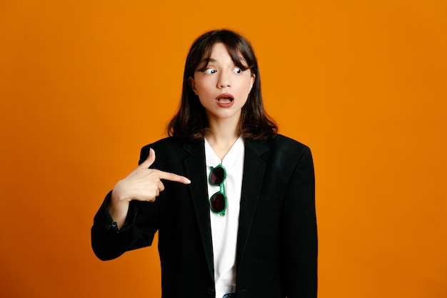 Surprised points at side young beautiful female wearing black jacket isolated on orange background