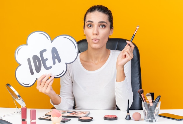 Surprised pretty caucasian woman sitting at table with makeup tools holding idea bubble and makeup brush isolated on orange wall with copy space