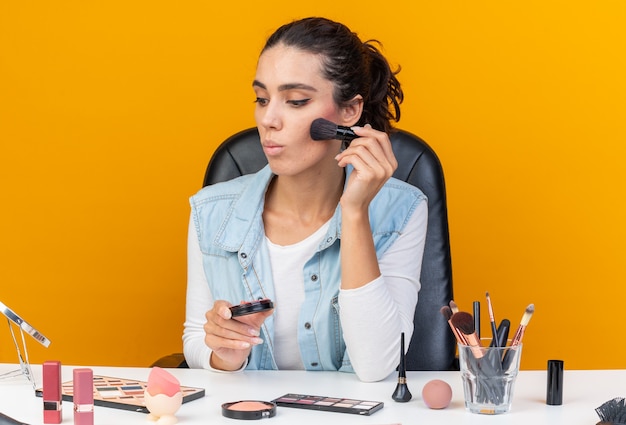 Surprised pretty caucasian woman sitting at table with makeup tools looking at mirror applying blush with makeup brush isolated on orange wall with copy space