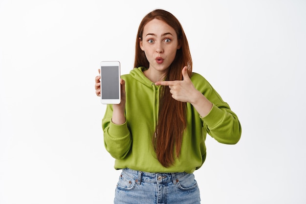 Surprised redhead woman pointing finger at smartphone app, gasping in awe, showing interesting application on mobile phone on white