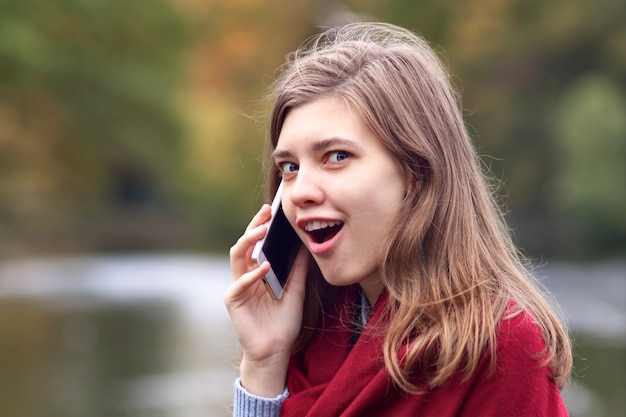 Surprised shocked happy young woman talking on cell mobile smart phone outdoors in autumn park with