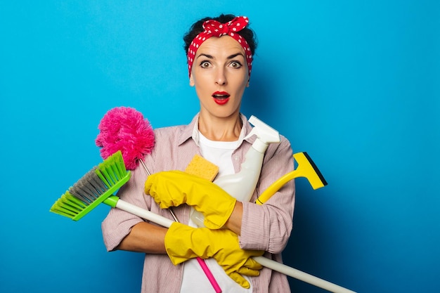 Surprised shocked young woman in yellow gloves holding cleaning accessories on blue background