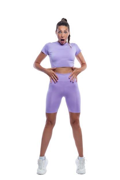 Surprised slender woman in purple sportswear points to her belly Sport active lifestyle beauty and health Isolated on white background Vertical Full height