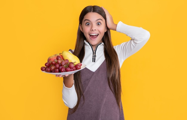 Surprised smiling child hold fresh fruit plate on yellow background vitamins