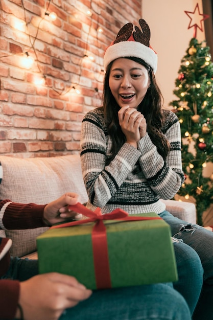 Surprised smiling woman and sister with Christmas gift box opening pulling red bow ribbon on boxing day. young girls celebrating xmas eve together with tree beside. asian lay excited holding hands.