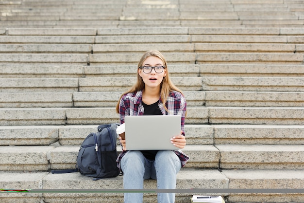 Surprised student girl sitting on stairs working with laptop, preparing for exams and drinking coffee outdoors, in university campus. Technology, education and remote working concept, copy space