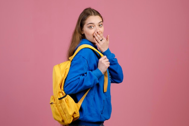 Surprised teen girl showing amazement by keeping hand on mouth while carrying backpack indoors