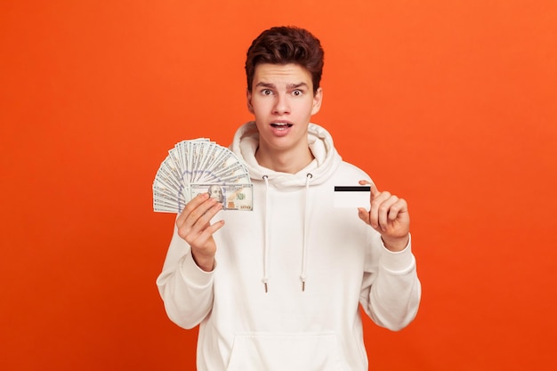 Surprised teenager in white casual style sweatshirt shocked with credit card cashback percentage holding dollars and card in hands. Indoor studio shot isolated on orange background