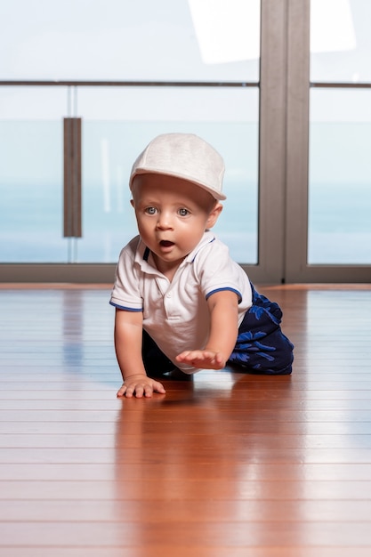 A surprised toddler boy in a fashionable cap with big blue eyes is crawling on the floor