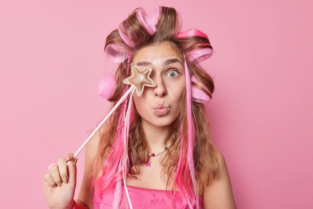 Surprised woman covers eye with magic wand keeps lips rounded applies hair rollers for making hairstyle wears dress isolated over pink background prepares for special occasion and celebration