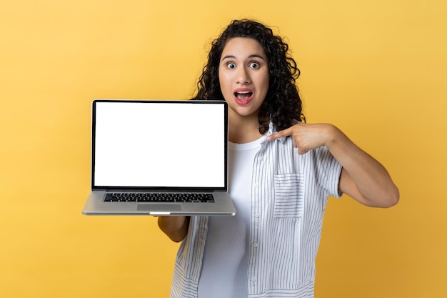 Surprised woman holding and pointing laptop with empty display copy space for advertisement