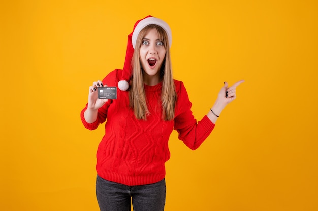 Surprised woman holding template credit card and pointing at copy space wearing santa hat