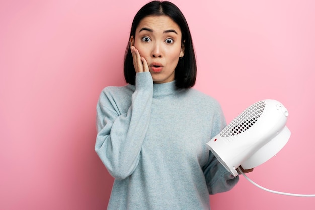 Surprised woman in warm clothes holding radiator on pink background