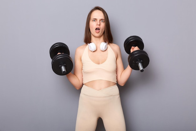 Surprised woman wearing top holding barbells in hands isolated on gray background doing sport exercising with heavy dumbbells screaming