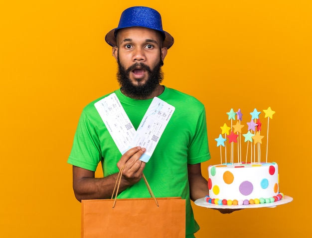 Surprised young afro-american guy wearing party hat holding gift bag and cake with tickets isolated on orange wall