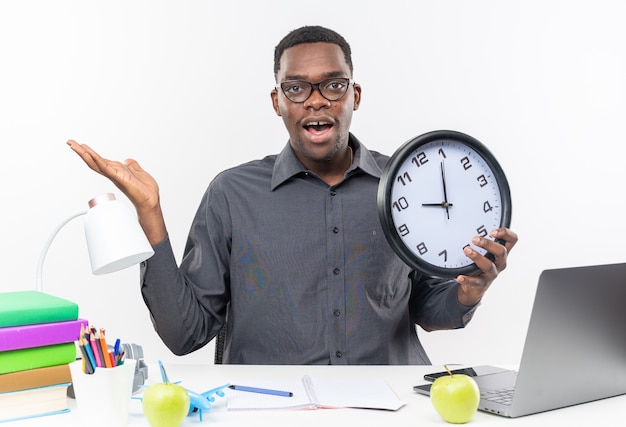 Surprised young afro-american student in optical glasses sitting at desk with school tools holding clock and keeping hand open isolated on white wall