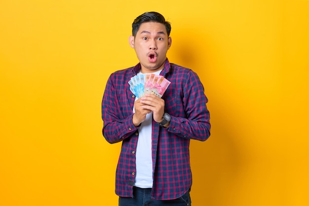 Surprised young Asian man in plaid shirt holding money banknotes with open mouth isolated on yellow background