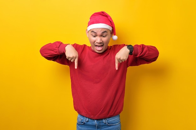 Surprised young Asian man in Santa hat pointing fingers down demonstrating free copy space for advertisement on yellow studio background celebration Christmas holiday and New Year concept