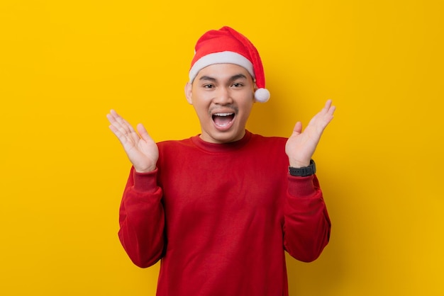 Surprised young Asian man in Santa hat raising hands and looking at camera with excitement expression on yellow studio background celebration Christmas holiday and New Year concept